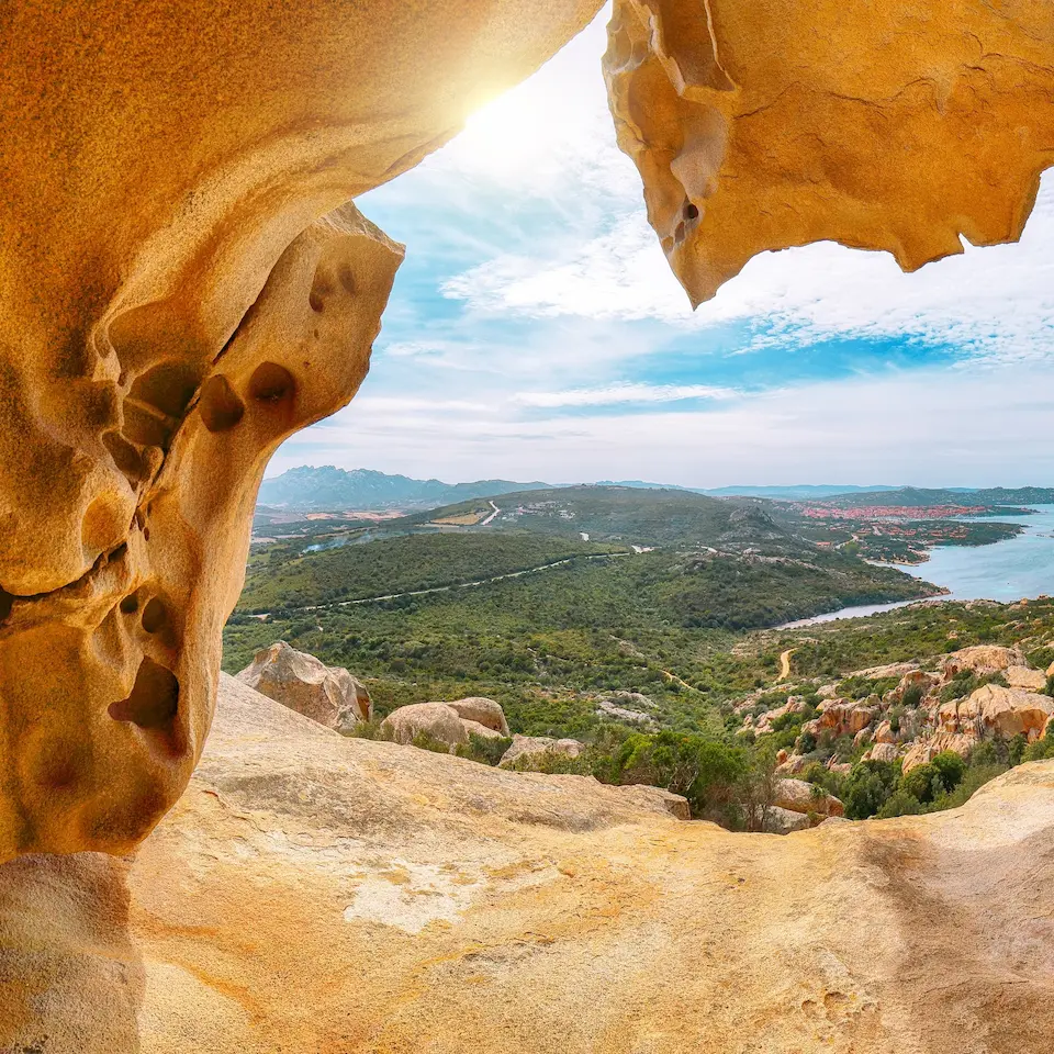 Ausblick durch eine natürliche Felsformation auf eine grüne Landschaft mit Hügeln, Wasser und Himmel