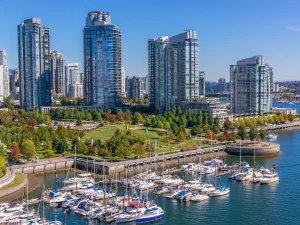 Das Bild zeigt eine beeindruckende Skyline von Vancouver, Kanada, mit modernen Wolkenkratzern, die entlang des Hafens aufragen. Im Vordergrund sind Yachten und Boote im Wasser zu sehen, während das grüne Gelände und Parks eine angenehme Balance zwischen Natur und urbanem Raum bieten. Der klare Himmel und die Sonne lassen die Szenerie noch lebendiger erscheinen. Vancouver ist bekannt für seine Kombination aus urbanem Leben und wunderschöner Natur.