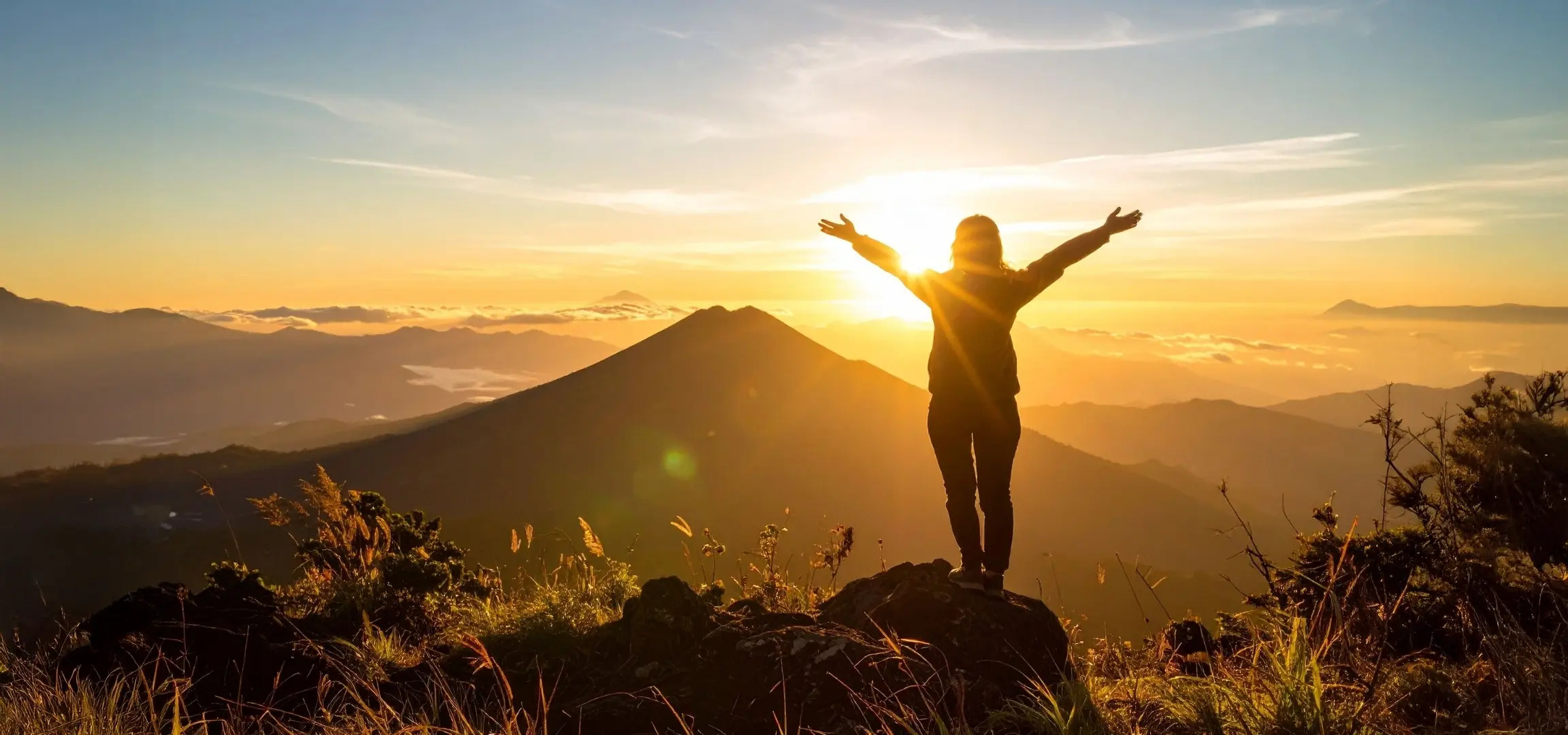 Person steht mit ausgebreiteten Armen auf einem Berggipfel im Sonnenaufgang mit Blick über eine weite Gebirgslandschaft