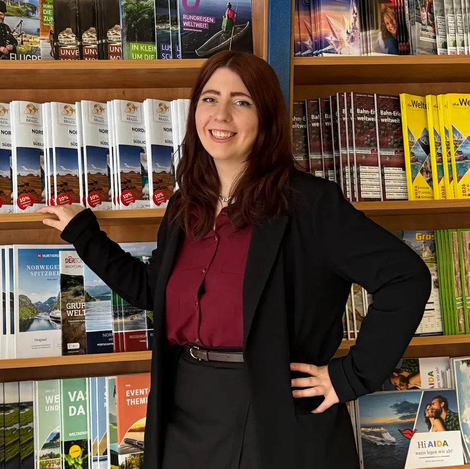 Tourismuskauffrau steht lächelnd vor einem Regal mit Reiseprospekten und Katalogen in einem Reisebüro