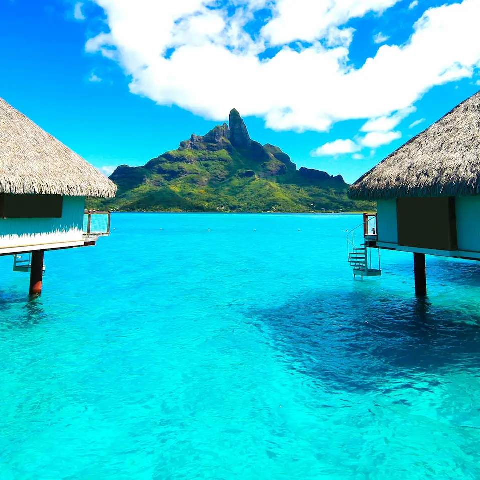 Überwasserbungalows stehen in einer türkisfarbenen Lagune, im Hintergrund eine grüne Insel unter blauem Himmel