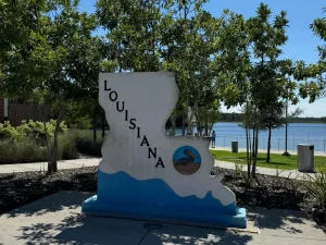 Auf dem Bild ist ein Denkmal in Form des US-Bundesstaates Louisiana zu sehen. Darauf steht der Name „Louisiana“ in schwarzen Buchstaben, und in der unteren rechten Ecke ist ein Kreis mit dem Symbol eines Pelikans – dem Staatsvogel von Louisiana – abgebildet. Im Hintergrund sieht man Bäume, eine Promenade sowie ein Gewässer, was die typische südliche, wasserreiche Landschaft des Bundesstaates widerspiegelt.